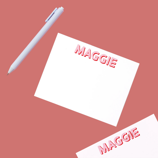 A white stylus pen and two white name tags printed with the name 'Maggie' in red 3D letters, arranged on a solid pink background.