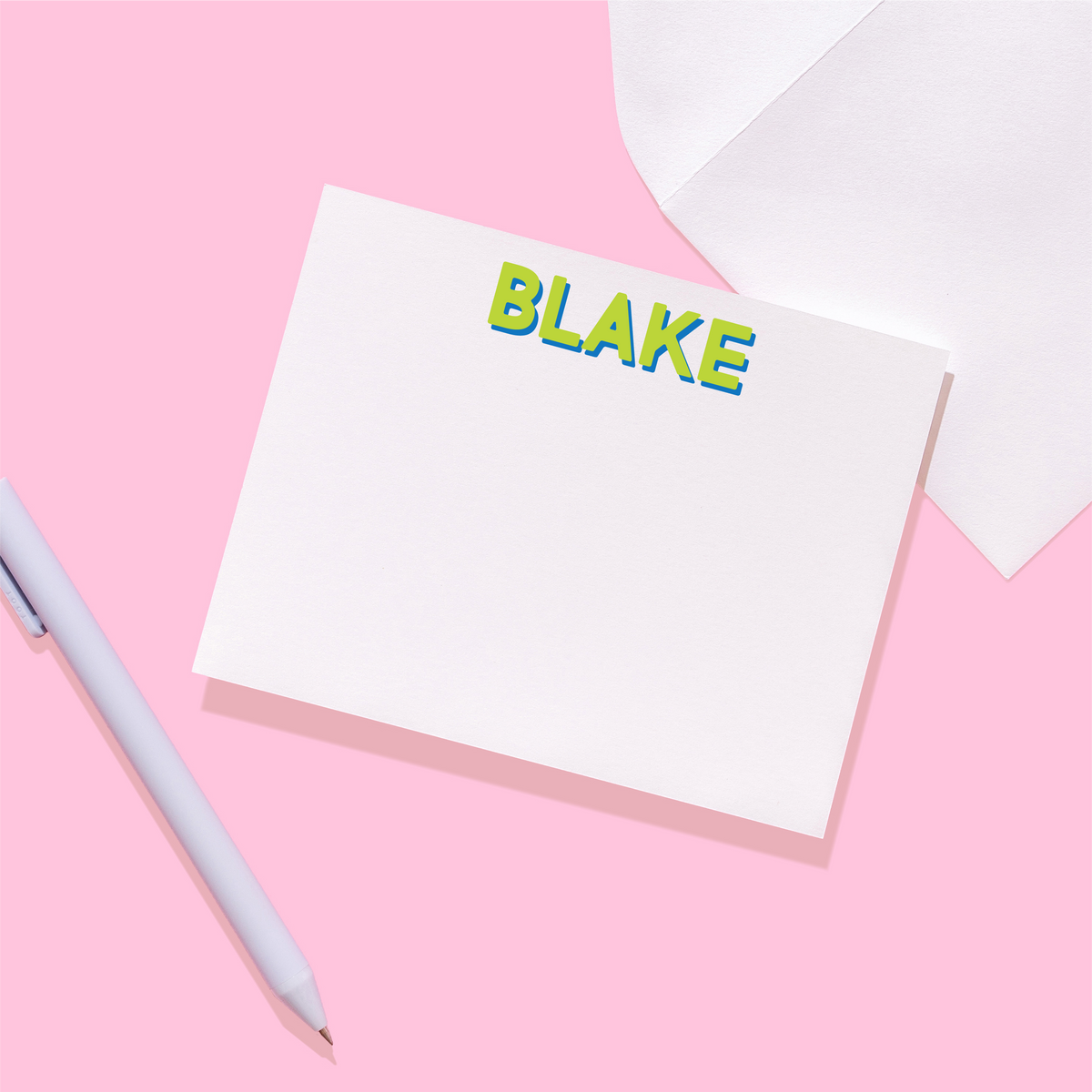 Note card with 'BLAKE' printed on it, placed on a pink surface with a pen and envelope.