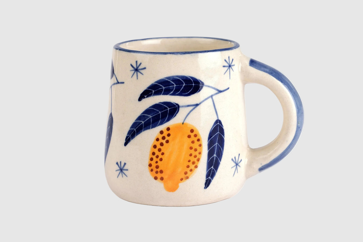 A ceramic mug with a cream-colored glaze, decorated with a blue and orange botanical pattern of leaves and a stylized fruit, shown on a solid white background.