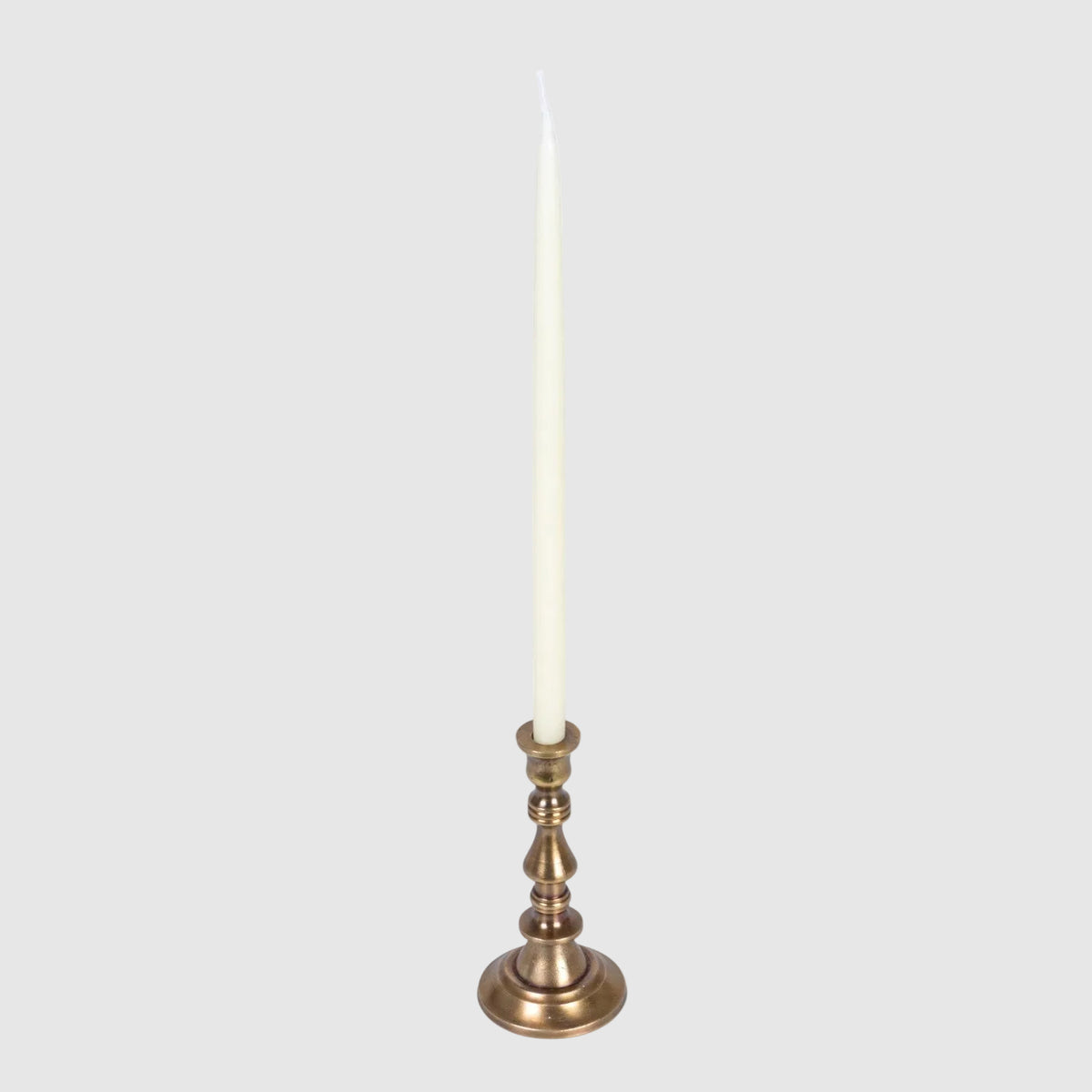 A single tall taper candle in an off-white wax finish, set in a polished brass candlestick holder with a flared round base, isolated on a white background.